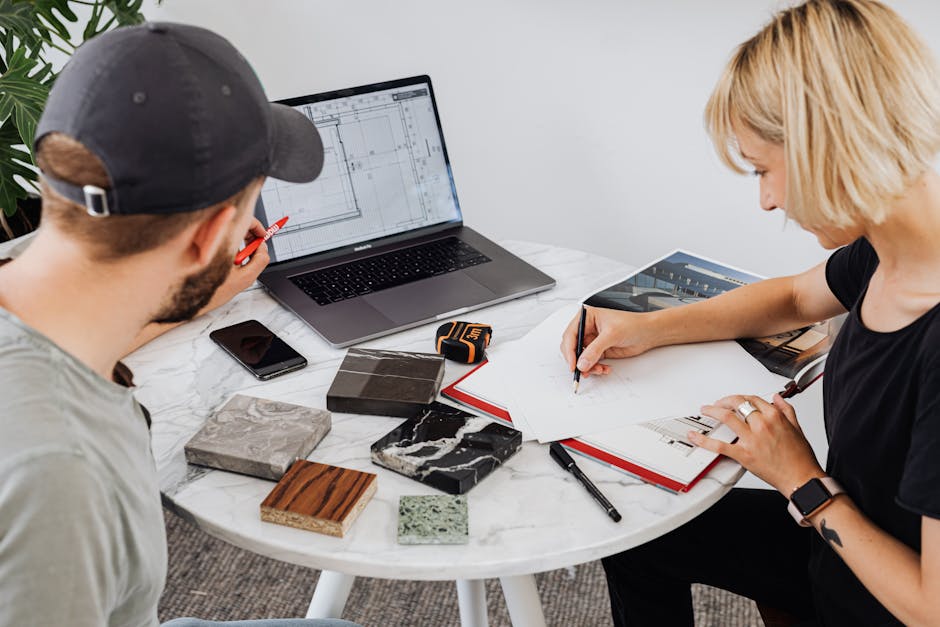 Two designers collaborating on a project, utilizing a laptop and drafting materials on a round table.