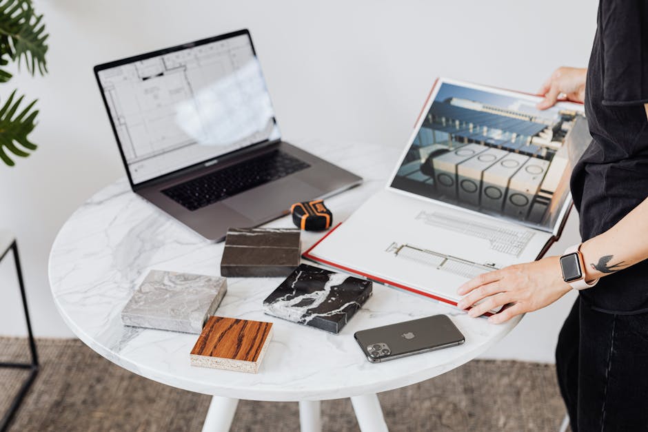 Architect reviews design plans on a marble table with laptop, phone, and material samples.
